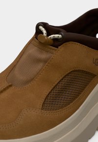 Brown suede slip-on shoe with mesh panels, textured surface, elastic lacing, and a rubber sole. Features subtle branding on the side.
