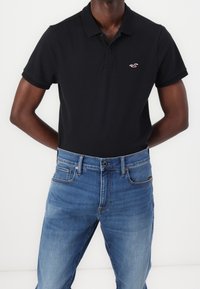 Polo de algodón negro con mangas cortas y cuello, con un pequeño logo bordado en el pecho, combinado con jeans de mezclilla azul.