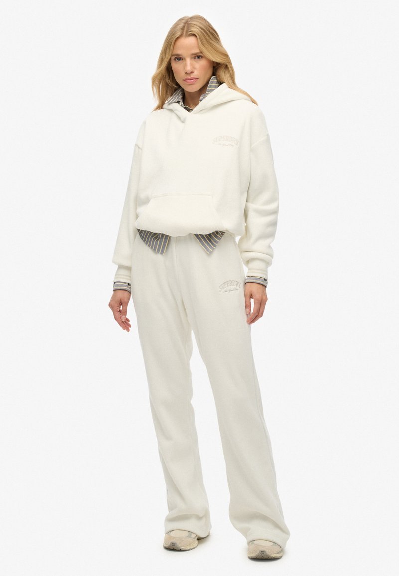 Mujer vistiendo una sudadera de gran tamaño color crema y pantalones de chándal a juego, con una camiseta de rayas debajo, de pie frente a un fondo blanco.