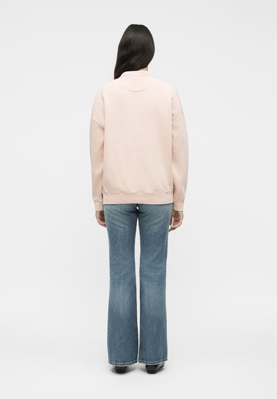 Femme aux longs cheveux foncés portant un sweat-shirt rose clair et un jean évasé bleu, debout, dos à un fond blanc uni.