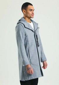 Grauer Kapuzenmantel mit langem Schnitt, ausgestattet mit einem Reißverschluss vorne, seitlichen Knöpfen und einem sportlichen Design. Hergestellt aus einem weichen, strukturierten Material.