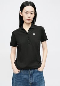 Črna polo majica z teksturirano obdelavo, ki ima ovratnik in tri gumbe na sprednji strani. Na prsih je viden majhen poudarek z logotipom.