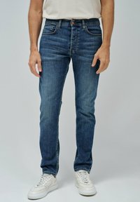 Jeans de mezclilla en azul oscuro con un ajuste Slim, diseño de cinco bolsillos, costuras en contraste y cierre de botón, combinados con zapatillas blancas.