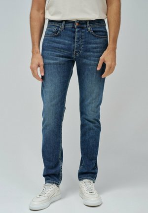 Jeans de mezclilla en azul oscuro con un ajuste Slim, diseño de cinco bolsillos, costuras en contraste y cierre de botón, combinados con zapatillas blancas.