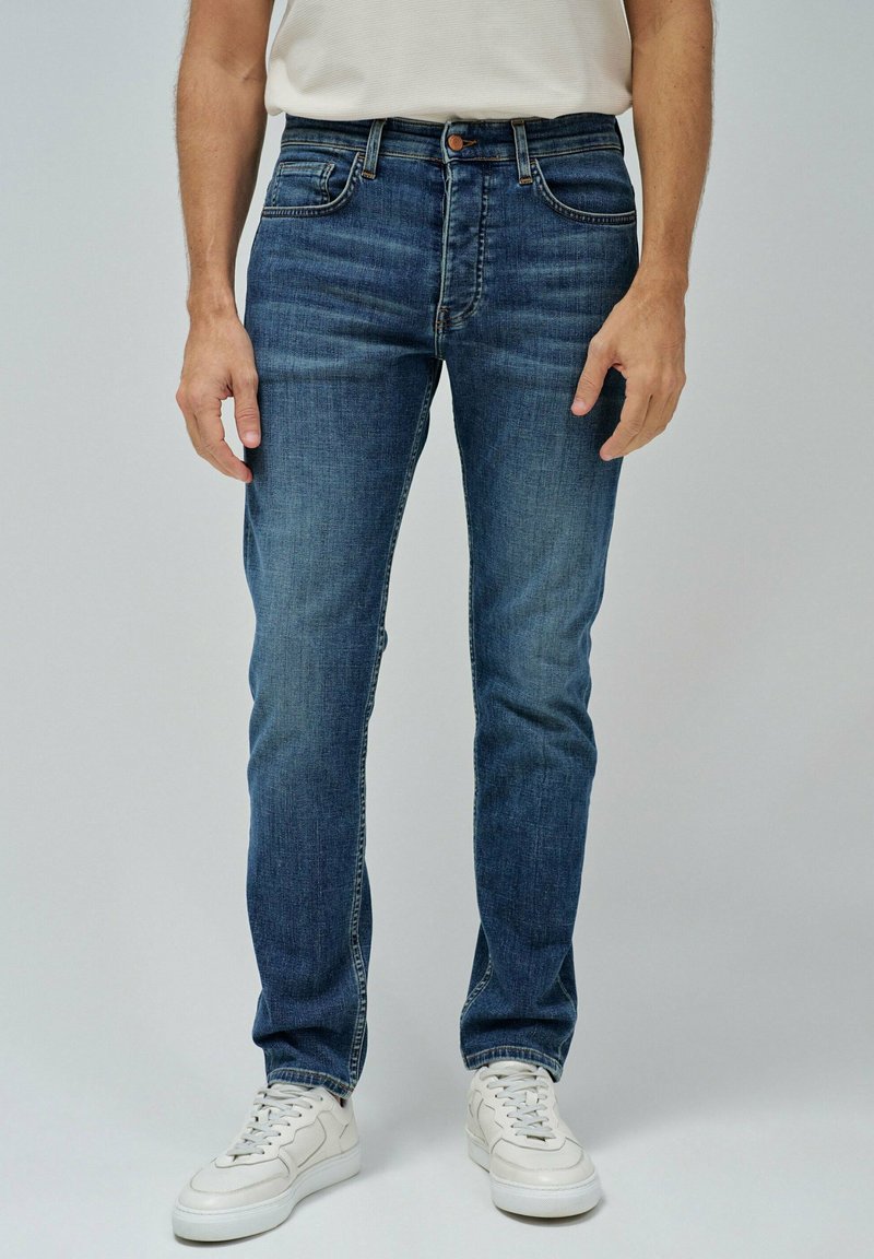 Jeans de mezclilla en azul oscuro con un ajuste Slim, diseño de cinco bolsillos, costuras en contraste y cierre de botón, combinados con zapatillas blancas.
