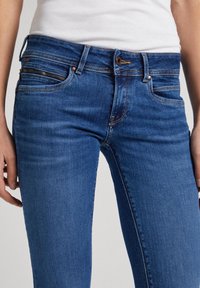 Blå jeans i denim med en figurnära design, detaljer med dragkedjefickor, kopparfärgade detaljer och en jämn textur.