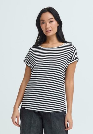 Kobieta o długich czarnych włosach ubrana w czarno-biały paskowany krótki rękaw i ciemne szerokie spodnie, stojąca na jasnym tle.