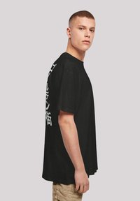 Black cotton t-shirt with short sleeves, featuring a loose fit and large white text on the left side, displayed on a model in profile.