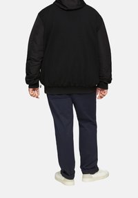 Schwarze gepolsterte Jacke mit strukturiertem Design, Kapuze und gerippten Bündchen, kombiniert mit navyblauen Hosen und weißen Sneakers. Ansicht von hinten.