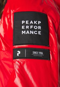 Rød dunjakke med en blank finish. Har et sort logoemblem med teksten "PEAK PERFORMANCE" og en etiket, der angiver "SINCE 1986, DESIGNED IN SWEDEN."