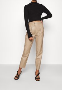 Haut noir à col roulé côtelé, associé à un pantalon taille haute en similicuir beige. Le mannequin porte des sandales noires à lanières, debout avec une main sur la hanche.