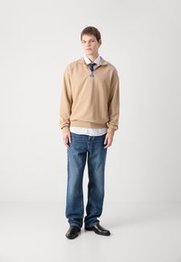 Felpa con cappuccio beige e colletto con zip, indossata sopra una camicia bianca, abbinata a jeans blu larghi e scarpe nere.