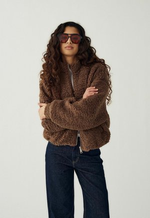 Ženska z dolgimi kodrastimi lasmi, ki nosi velika rjava sončna očala, rjavo teksturirano bunda z zadrgo in temno modre jeans hlače, z rokami prekrižanimi.