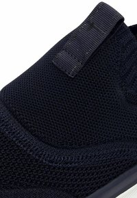 Scarpa slip-on lavorata a maglia di colore blu scuro con tessuto testurizzato, cinturino elastico e suola bianca robusta, con cuciture visibili e design senza cuciture.