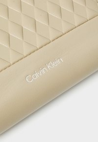 Beżowy portfel z pikowanym diamentowym wzorem oraz gładkimi skórzanymi akcentami. "Calvin Klein" jest wytłoczone na biało w dolnym prawym rogu.