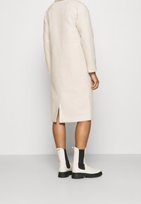 Cappotto lungo color crema, simile alla lana, con spacco posteriore, abbinato a stivaletti bianchi con pannelli laterali neri e suola robusta.