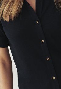 Camisa de botón de manga corta azul marino con escote en V y botones de madera. La tela parece suave con un ligero brillo.
