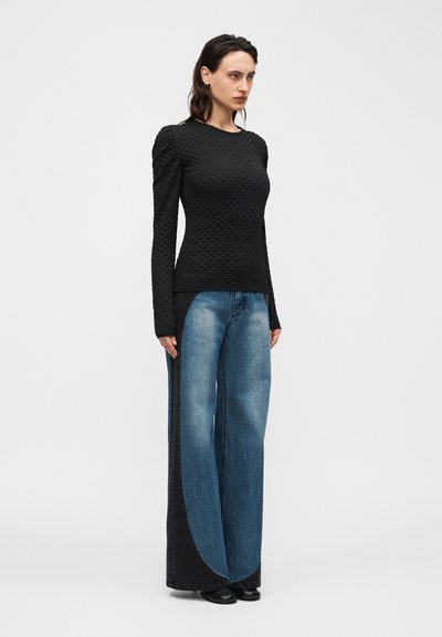 Γυναίκα που φοράει γυαλιστερό μαύρο τοπ με υφή και μακριά μανίκια με φουσκωτούς ώμους και τζιν παντελόνι με φαρδιά πόδια και σκούρα πλαϊνά πάνελ, σε μονόχρωμο φόντο.