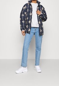 Niebieska kurtka puchowa w kremowy geometryczny wzór, zestawiona z szarym swetrem, jasnoniebieskimi dżinsami i białymi trampkami.