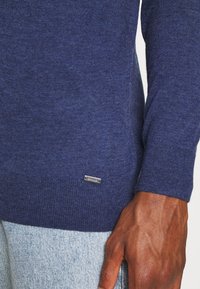 Navy sweater made of textured fabric, featuring a silver logo tag near the hem and ribbed cuffs, paired with light denim.