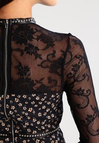 Schwarzes Kleid aus floraler Spitze mit einem Reißverschluss hinten, durchsichtigen langen Ärmeln und einem enganliegenden Oberteil mit kleinem floralen Muster und Nietenakzenten.