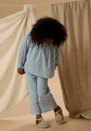 Dijete s kovrčavom kosom koje nosi svijetloplavu bluzu s izrezbarenom tkaninom i hlače, gleda dolje, stoji između bež i krem zastora od tkanine.