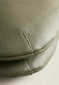 Grüne Ledertasche mit strukturierter Oberfläche, kontrastierenden Nähten und abgerundeter Form. Auffälliges Nahtdetail betont das Design.