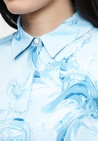 Κοντινό πλάνο ενός ατόμου που φορά ένα ανοιχτό γαλάζιο πουκάμισο με γιακά και κουμπί, με σπειροειδή μοτίβα που θυμίζουν νερό σε πιο σκούρες μπλε αποχρώσεις.