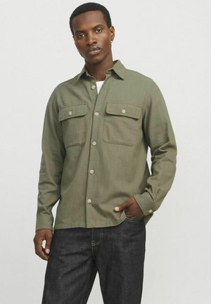 Olijfgroene longsleeve shirt met twee borstzakken, knoopsluiting en een relaxte pasvorm. Draagt over een wit T-shirt, gecombineerd met donkere jeans.