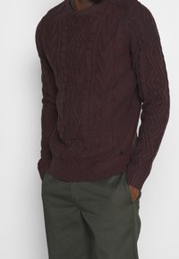 Dunkelburgunderfarbener Strickpullover aus Wolle, mit Rundhalsausschnitt, gerippten Bündchen und Saum, sowie strukturierten Mustern und einem kleinen Logo-Detail.