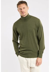 Homme portant un pull en col roulé vert olive et un pantalon écru, se tenant contre un fond blanc uni avec les mains dans les poches.