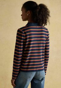 Dlhý rukáv, námornícky polo s červenými a olivovými pruhmi, s golierom a rovnými okrajmi. V kombinácii s džínsami s vysokým pásom v modrej farbe.