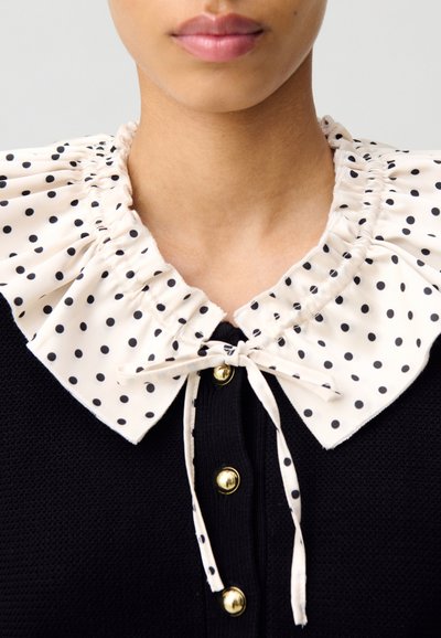 Jersey negro con un cuello fruncido de lunares crema. Presenta un lazo atado y botones dorados a lo largo del frente.