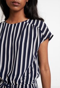 Blouse rayée bleu marine et blanc à manches courtes, avec des détails de épaules froncées et une taille cintrée. Tissu léger avec une texture lisse.