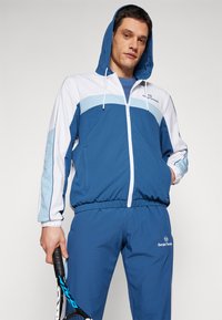 Blå treningsdrakt laget av lett materiale som har en zip-up jakke med hette, hvite detaljer og logo. Buksene har elastisk midje.