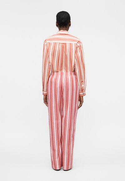 Άτομο με σκούρα μαλλιά σε κότσο, φοράει μακρυμάνικο πουκάμισο με γιακά και ψηλόμεσο παντελόνι με κάθετες κόκκινες και λευκές ρίγες, στέκεται με την πλάτη γυρισμένη.