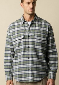 Uomo che indossa una camicia a quadri verde, navy e bianca con tasche a pattina sul petto sopra una maglietta bianca, in piedi contro uno sfondo beige.