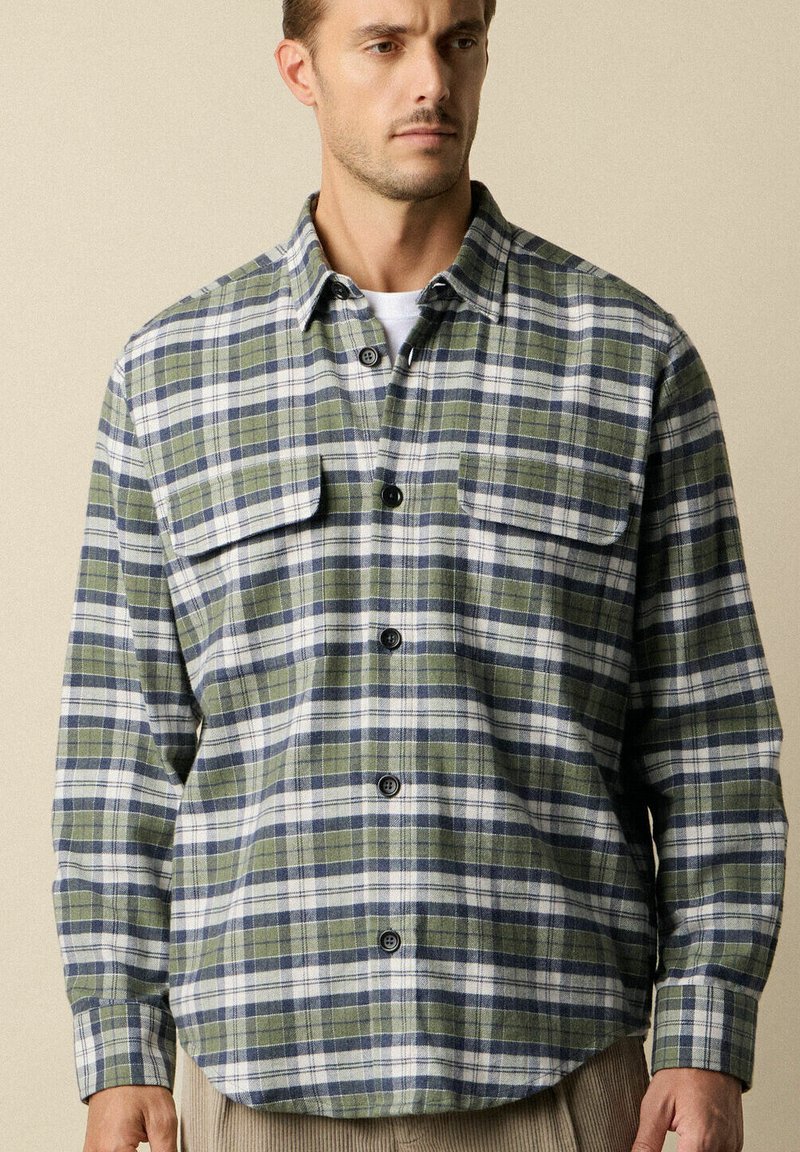 Uomo che indossa una camicia a quadri verde, navy e bianca con tasche a pattina sul petto sopra una maglietta bianca, in piedi contro uno sfondo beige.