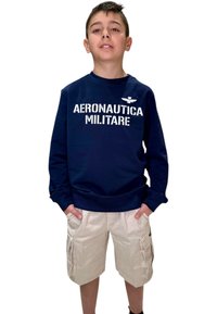 Felpa navy con la scritta "AERONAUTICA MILITARE" stampata in bianco, abbinata a pantaloni cargo beige con tasche laterali e vestibilità rilassata.