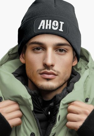 Black knit beanie with "AHOI" and anchor graphic. Model wears a green puffer jacket with black accents, showcasing layered textures.