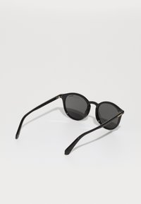 Ochelari de soare rotunzi, de culoare neagră, cu lentile întunecate, ramă subțire din plastic negru și brațe elegante, negre, cu detalii discrete ale mărcii.