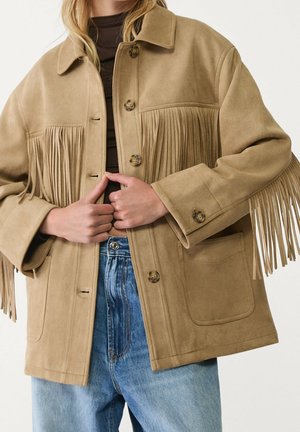 Personne portant une veste en daim beige avec fermeture par boutons et détails à franges sur la poitrine et les manches, associée à un jean bleu et un haut foncé.
