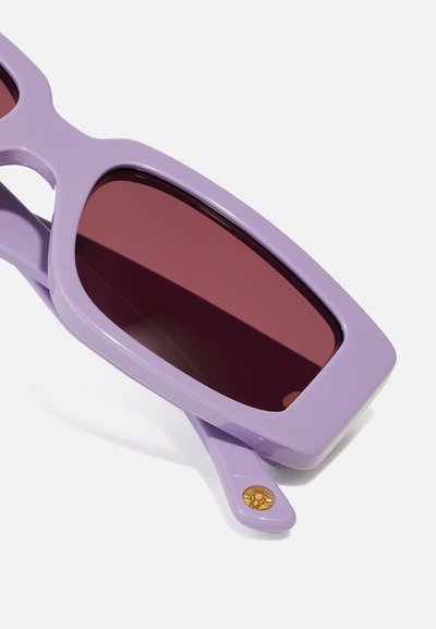 Lunettes de soleil violettes avec une monture rectangulaire épaisse et des verres rouge foncé. Un emblème solaire doré sur la branche ajoute un détail à la conception élégante.