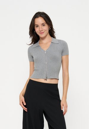 Jeune femme aux cheveux foncés portant un crop top gris côtelé à manches courtes avec fermeture éclair et un pantalon noir, souriante avec une main sur la hanche.
