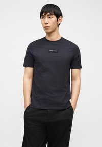 Νέος άνδρας που φοράει στενή μαύρη μπλούζα Armani Exchange με λογότυπο σε πατς στο στήθος, στέκεται με τα χέρια στις τσέπες μπροστά σε λευκό φόντο.