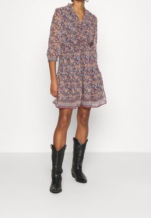 Frau trägt ein knielanges, geblümtes Kleid mit dreiviertellangen Ärmeln und schwarze Leder-Cowboystiefel, steht vor einem einfarbigen weißen Hintergrund.