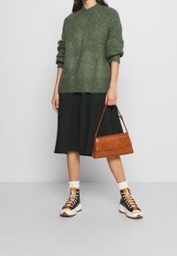 Maglione a maglia verde oliva, gonna midi nera, borsa in pelle effetto coccodrillo marrone e sneakers alte nere con accenti arancioni.