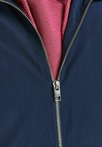 Marineblauwe jas met een zilveren rits, voorzien van een roze binnenlaag. De stof lijkt glad met een matte afwerking en elastische accenten.