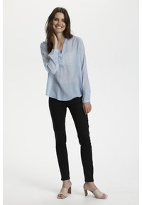 Blouse bleu clair à manches longues avec un motif géométrique, décolleté en v ; associée à un pantalon noir ajusté et à des escarpins beiges à bout ouvert.