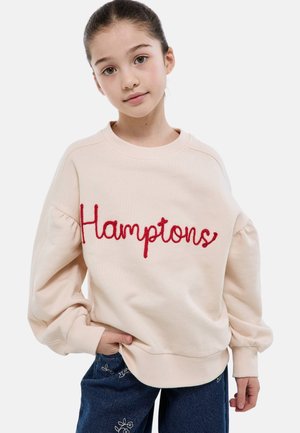 Jeune fille aux cheveux bruns portant un sweat beige avec l'inscription "Hamptons" en écriture rouge, debout avec une main dans la poche et une expression neutre.